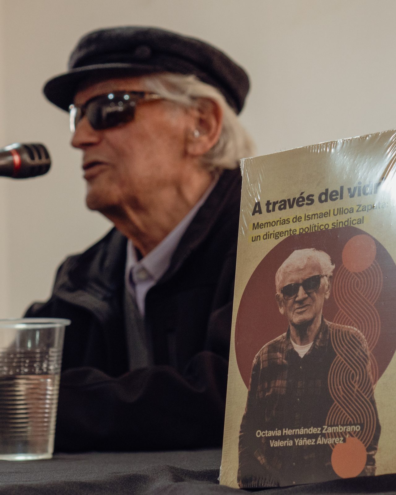 Ismael Ulloa Zapata habla frente al micrófono en el lanzamiento de sus memorias, con un ejemplar de «A través del vidrio» en pie sobre la mesa, a su lado.