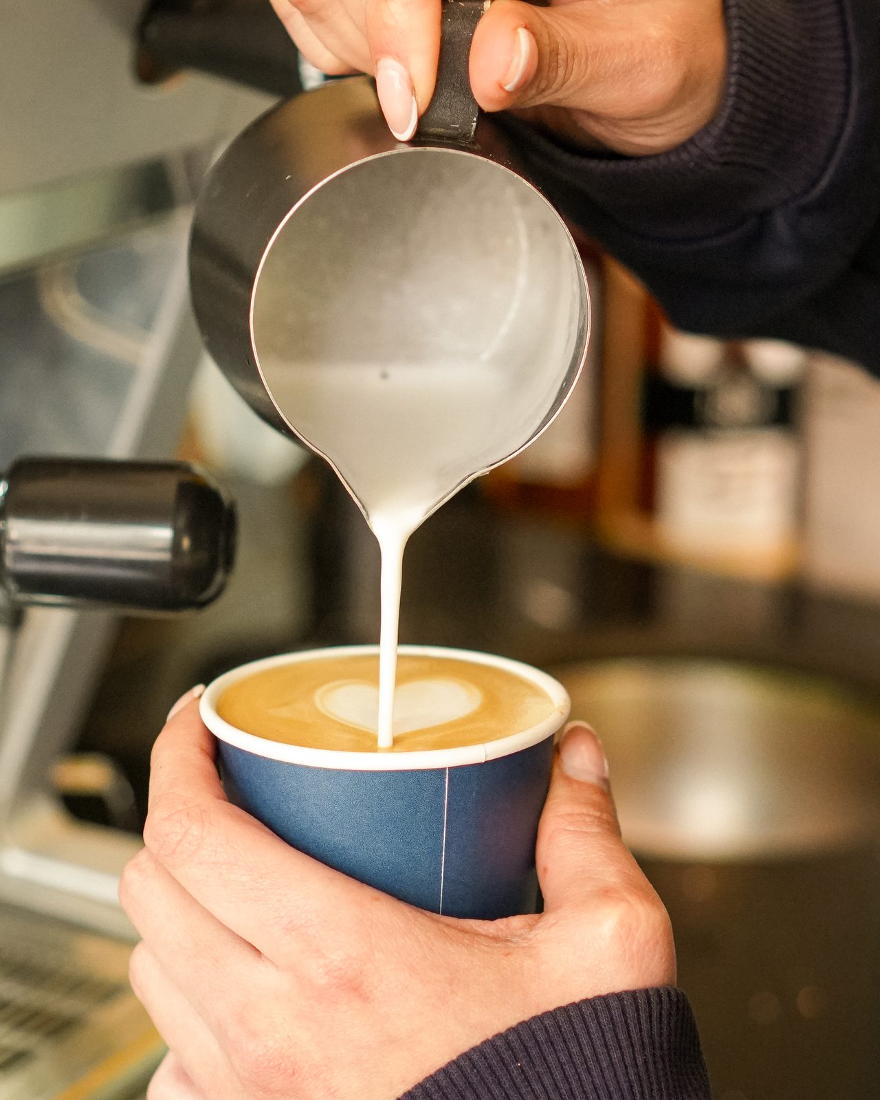 Manos vertiendo leche desde una jarra sobre un vaso de café, formando un corazón en la superficie, junto a la máquina de la barra.
